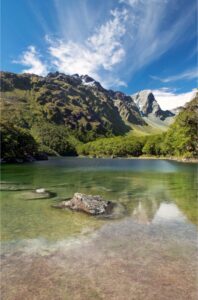 A clear mountain pool surround by vegetation-covered mountains under a blue sky in New Zealand on a Great Walk 